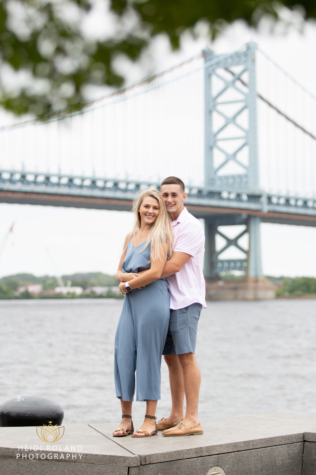 Newly engaged couple wrapping their arms around each other in front of Ben Franklin Bridge by Heidi Roland Photography