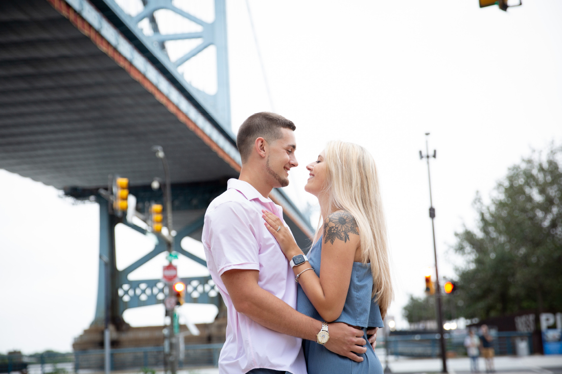 Engaged couple hugging and smiling near the Ben Franklin Bridge in Philly